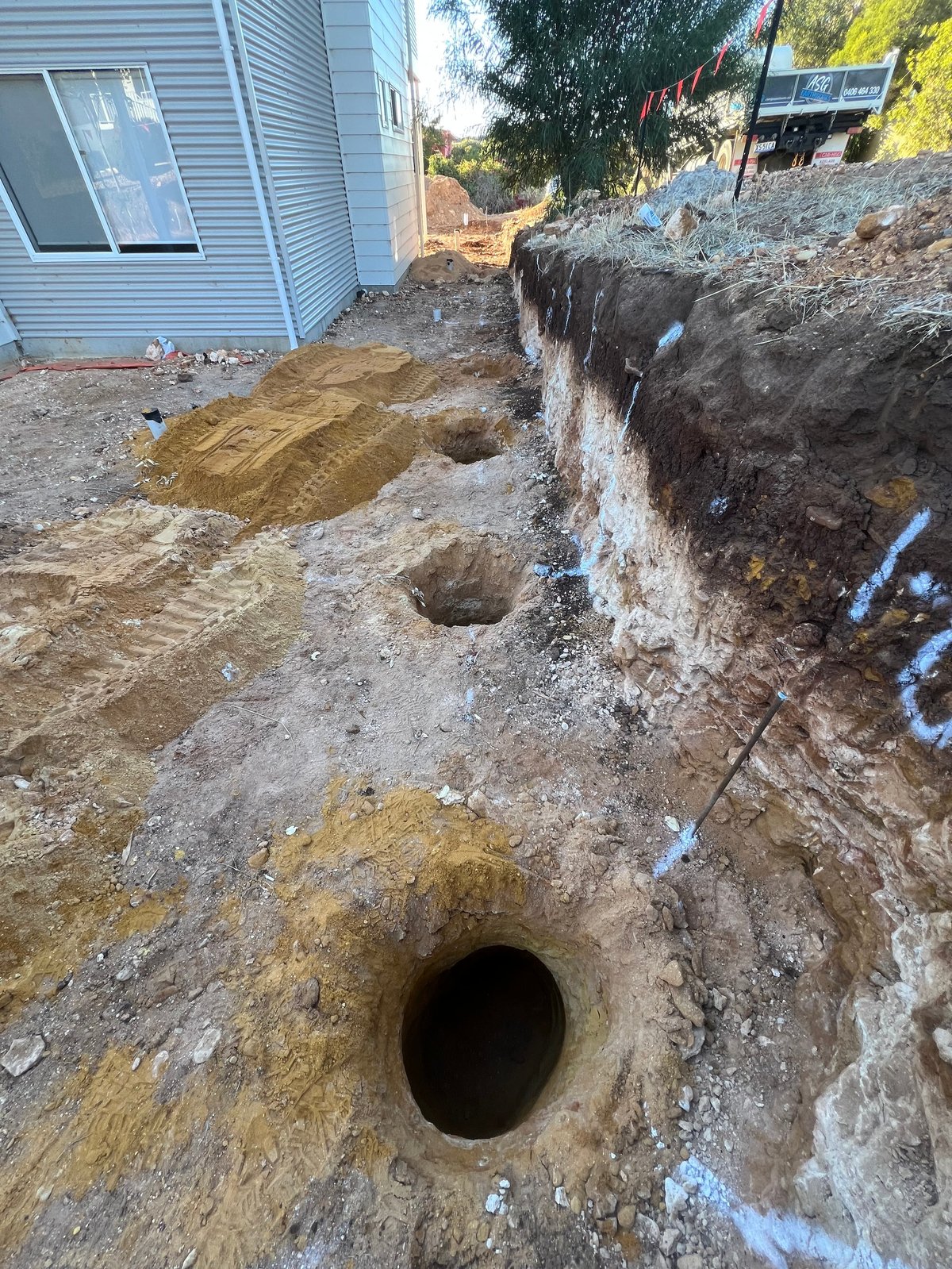 Excavated hole in dirt ground next to light blue mobile home during construction or utility work