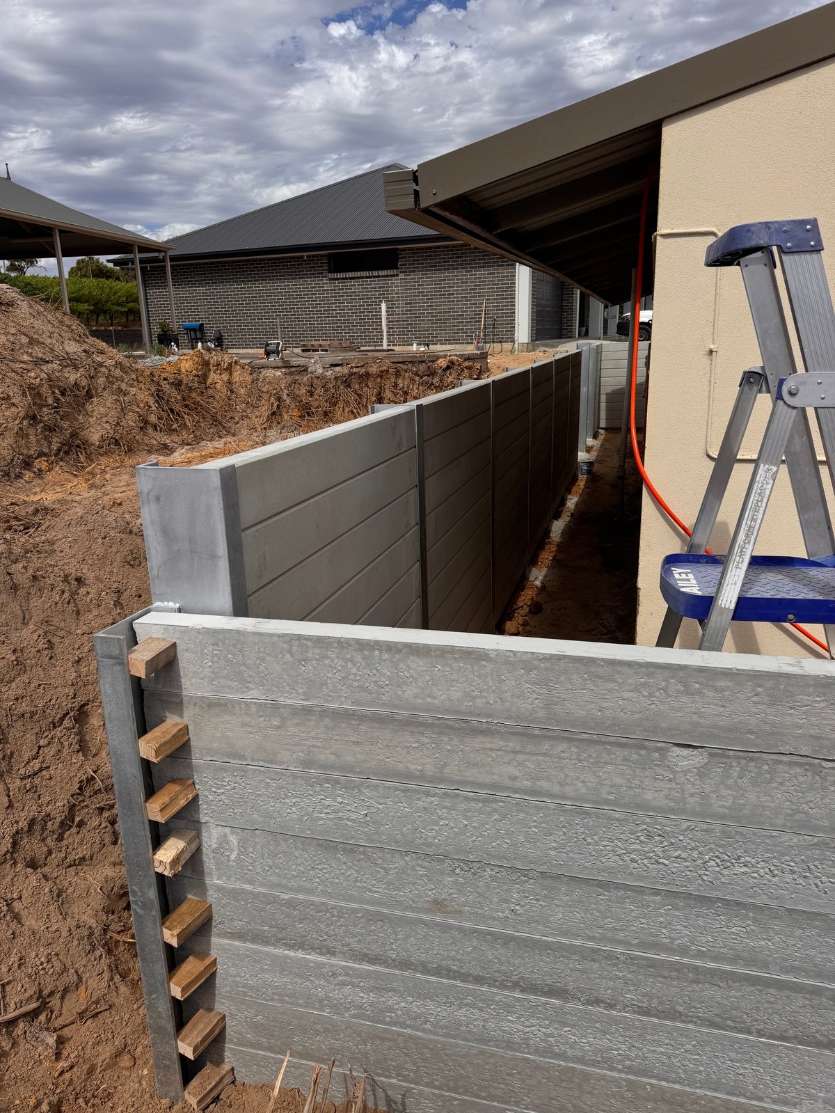 Construction site showing metal retaining walls and excavated foundation next to a modern brick building with a blue ladder visible
