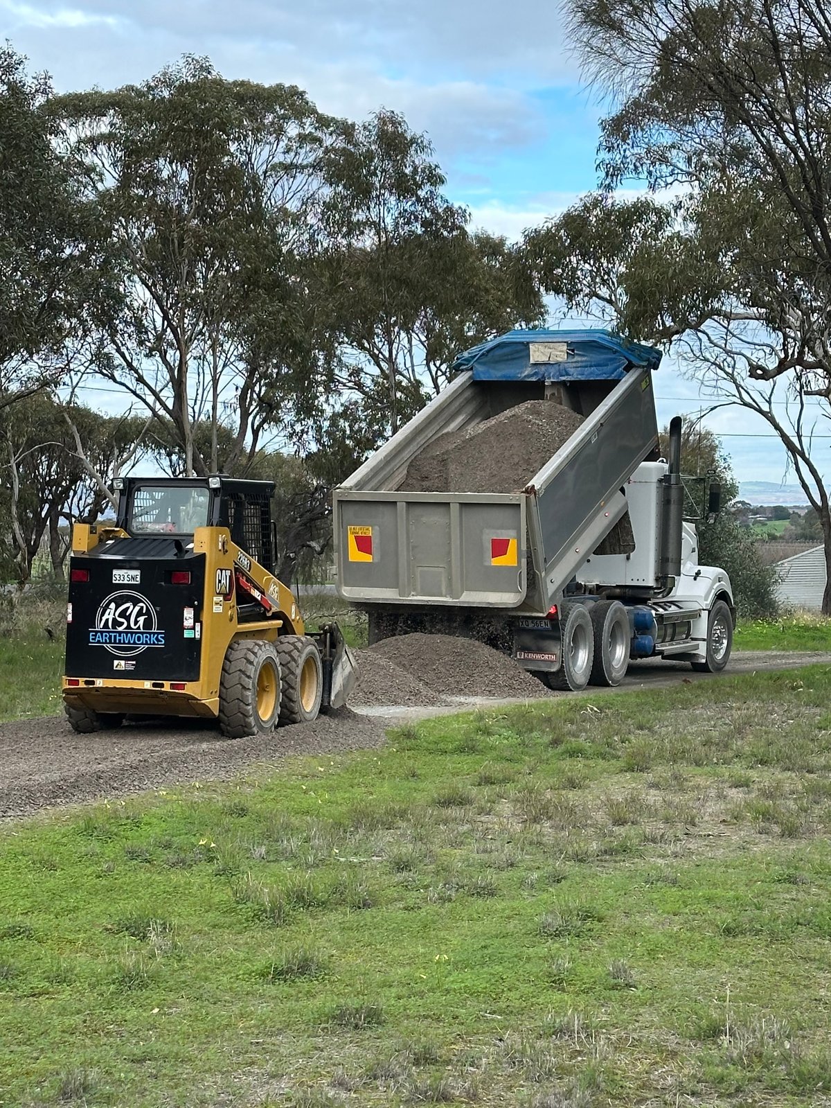 Yellow skid steer loader dumping gravel into a large dump truck on a rural road lined with eucalyptus trees