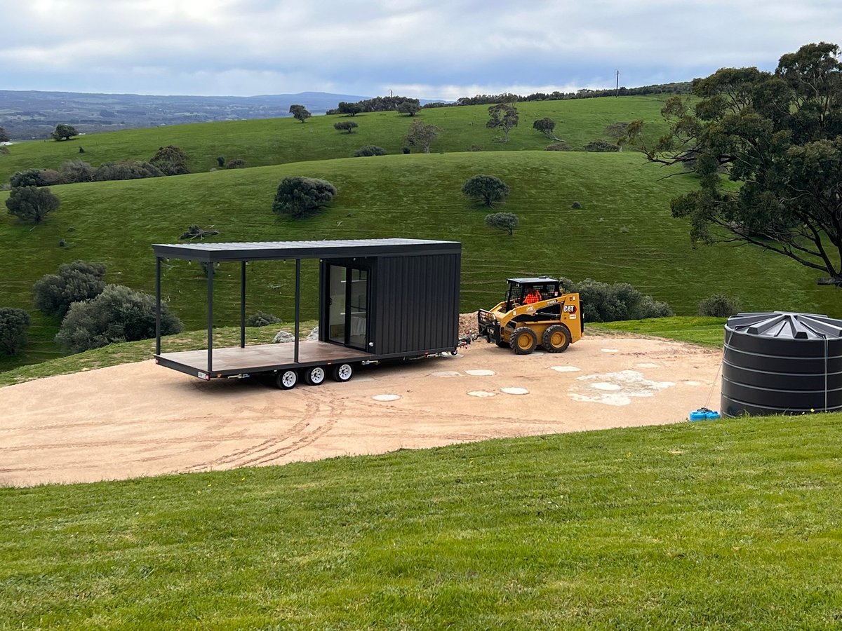 Modern black shipping container converted into a mobile home with covered porch on a cleared dirt pad in rolling green pastureland, with a yellow skid steer loader and water tank nearby