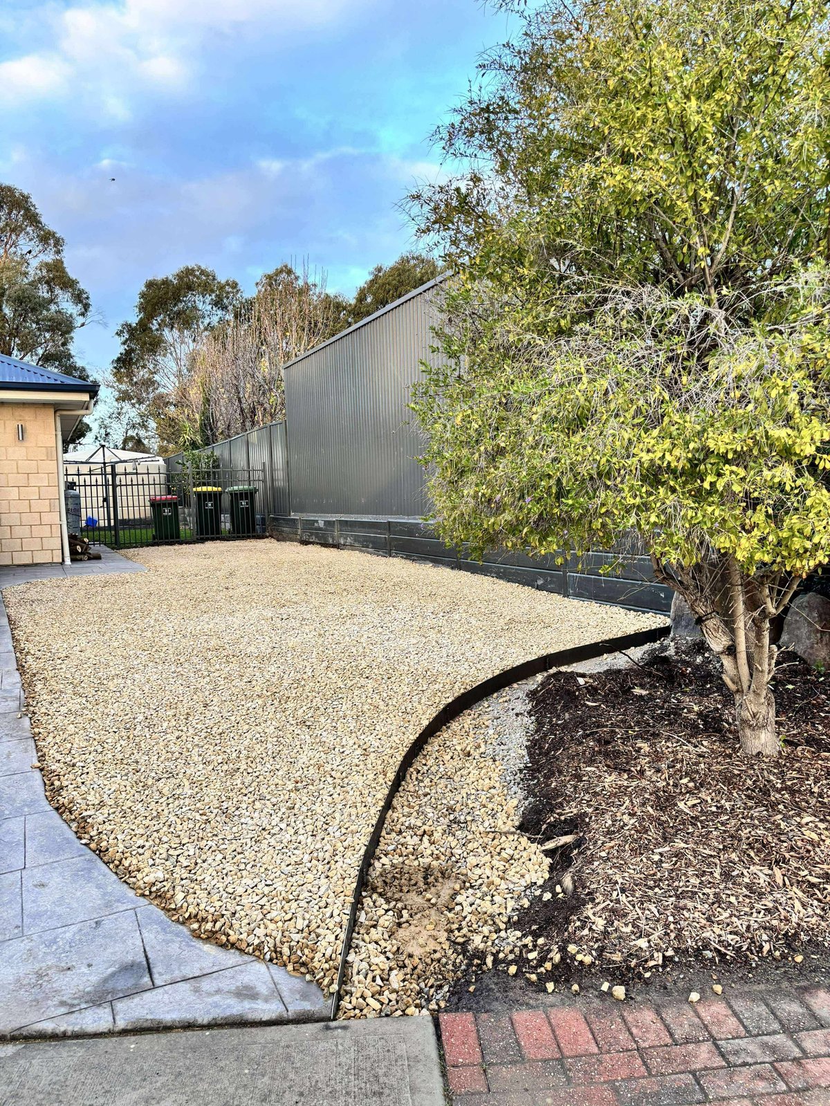 Modern backyard with gravel patio, dark corrugated metal fence, tree, and landscaping under blue sky