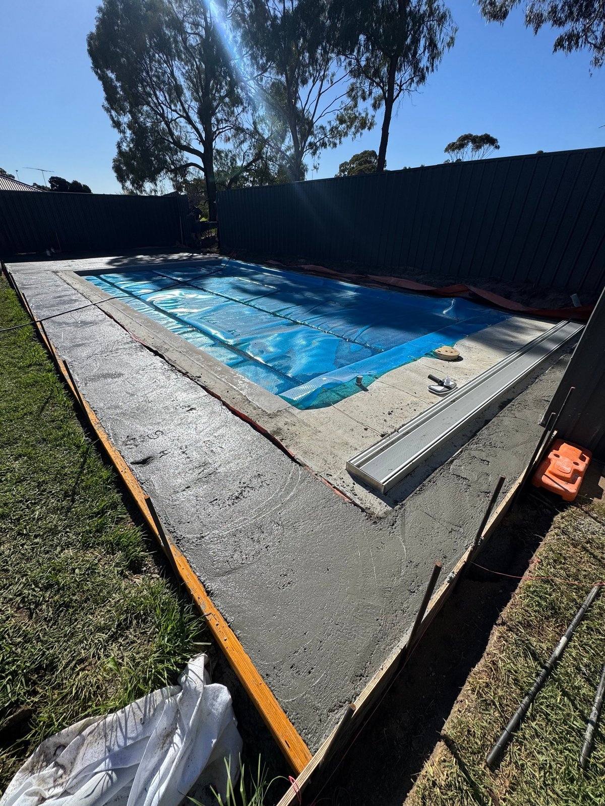 Residential swimming pool with blue cover surrounded by concrete deck, grass, and black fence on sunny day