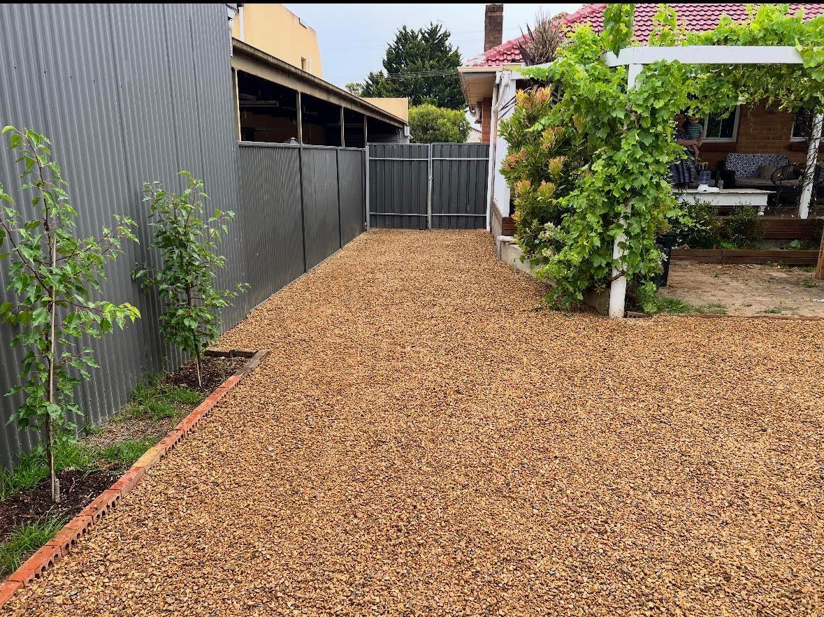 Paved backyard with gravel surface, metal fencing, storage structures, and climbing vines on wooden pergola