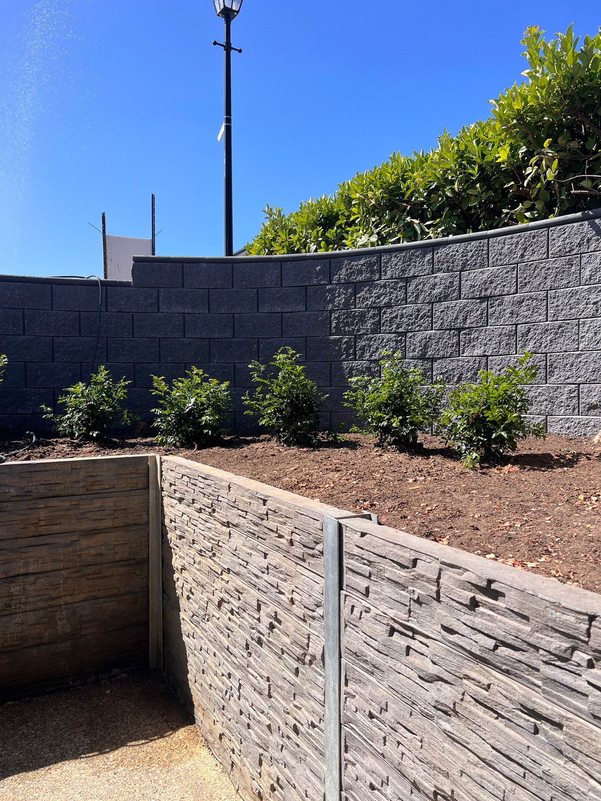 Backyard with raised wooden planter boxes, concrete block walls, shrubs, and a black street lamp against a clear blue sky
